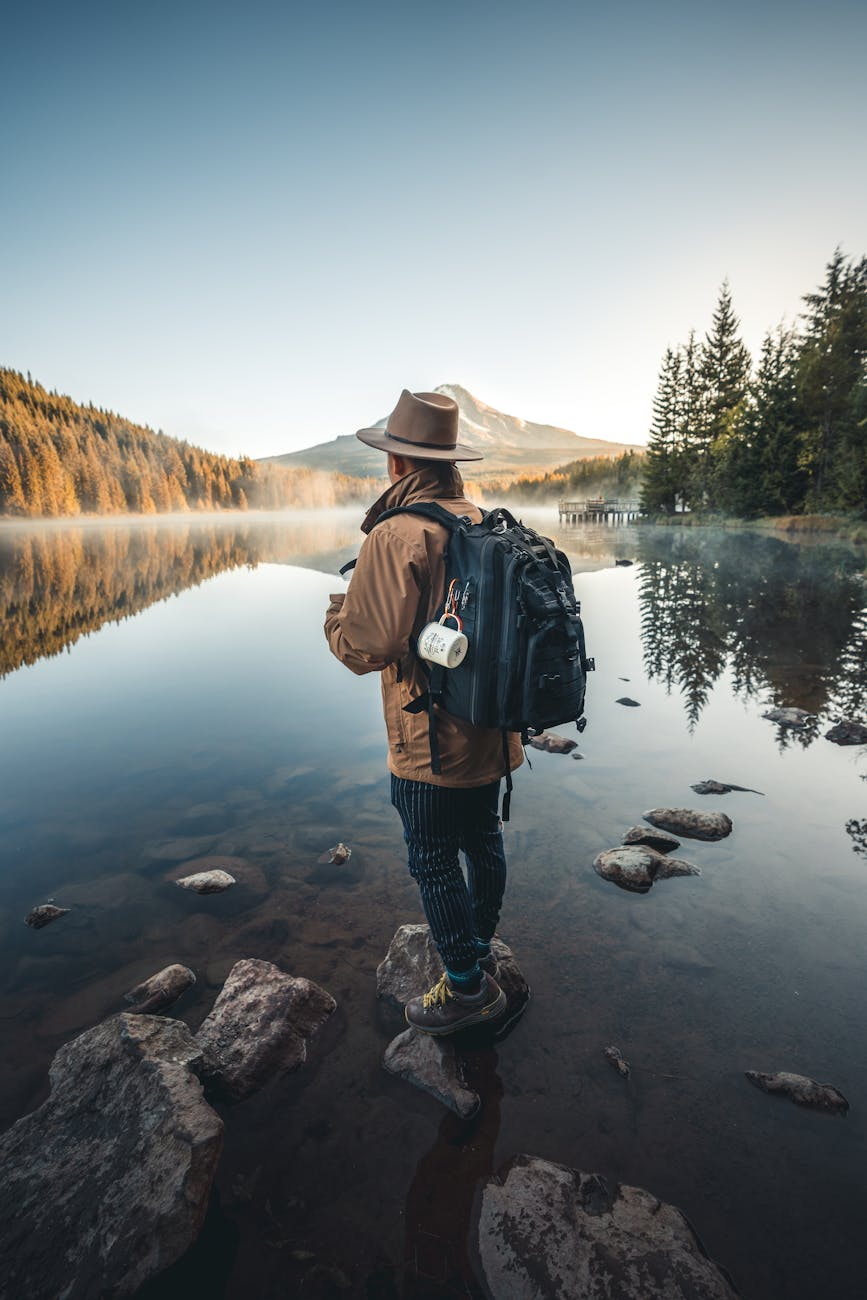 man in brown jacket and brown hat standing on rock near lake